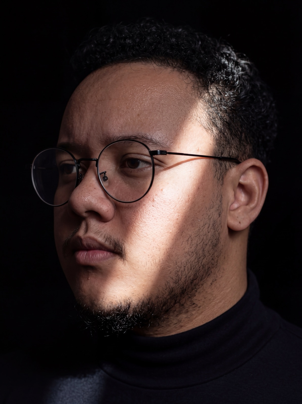 Retrato do Rodrigo Miguel, de óculos e blusa preta, com luz lateral e fundo escuro
